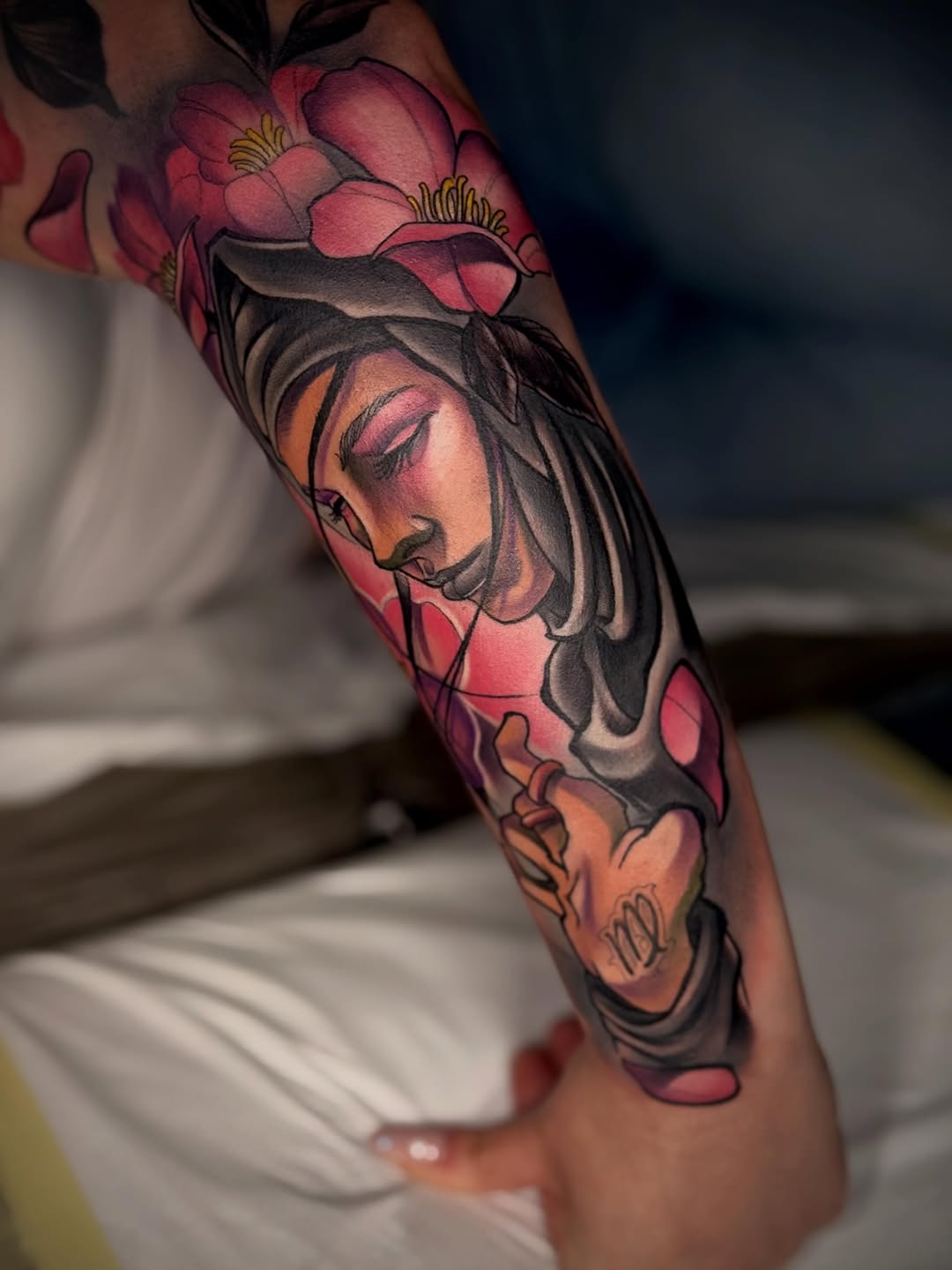 Colorful arm tattoo featuring a woman's face with closed eyes, pink and red flowers above, and orange accents, displayed on pale skin.