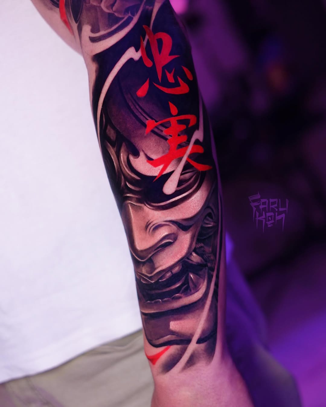 Colorful arm sleeve tattoo featuring stacked demon masks with red kanji characters and ornamental details, photographed under purple lighting.
