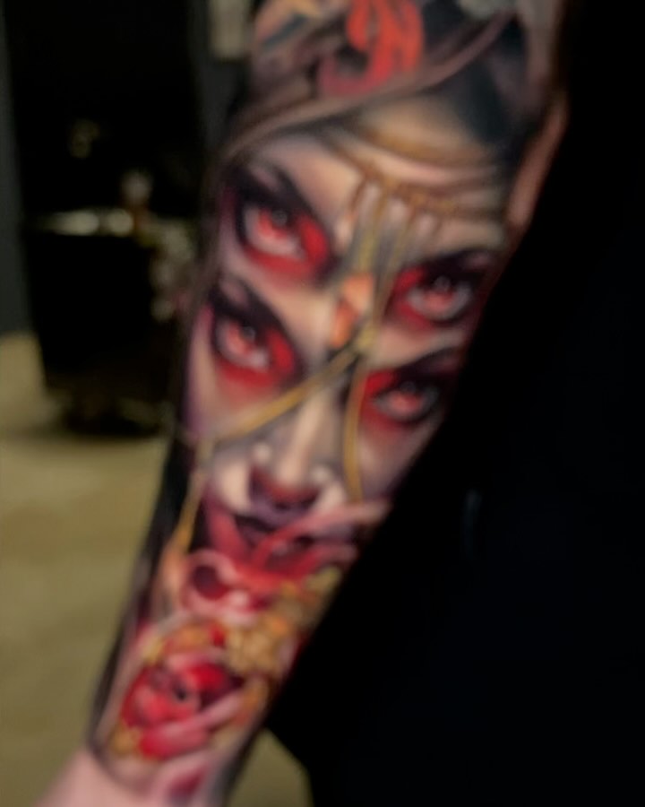 Colorful sleeve tattoo featuring multiple female faces with red eyes and flowers in shades of red, gold, and purple on the forearm and upper arm.