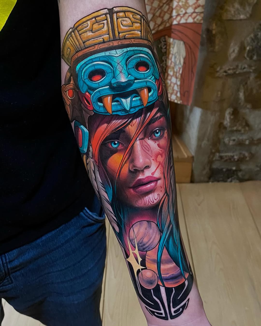 Colorful forearm tattoo featuring a blue tribal mask above a woman's face with blue eyes and red-orange facial markings, surrounded by geometric patterns.