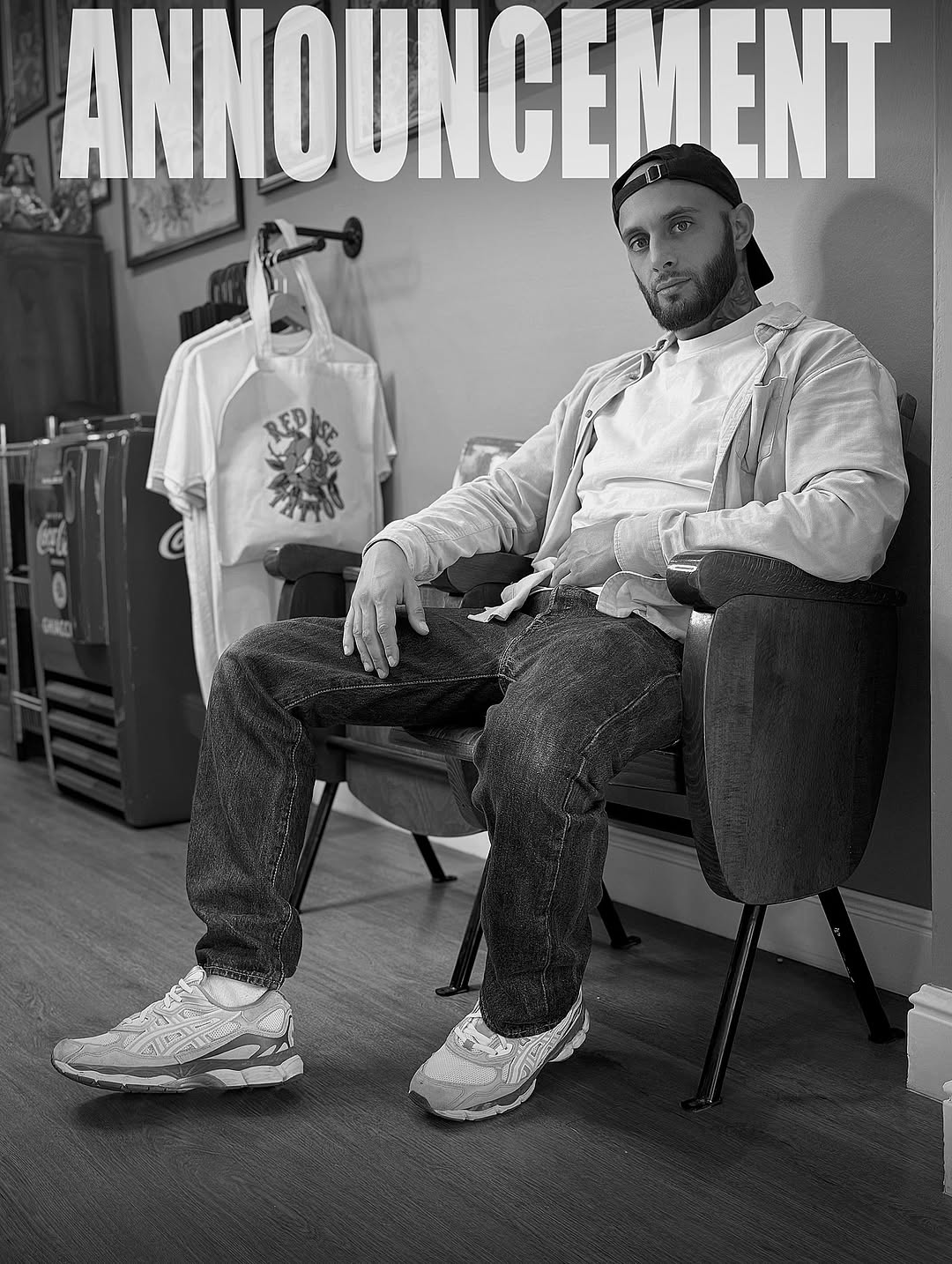 Bearded man wearing white jacket and cap sits in chair displaying arm tattoo with dark ink design on upper arm, studio setting with merchandise visible.