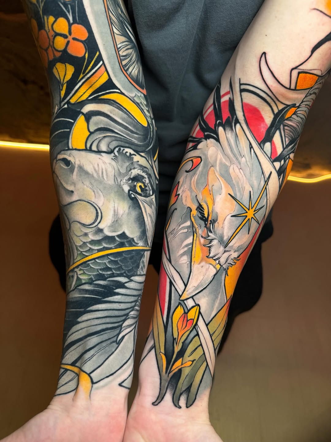 Two forearms display matching animal head tattoos with orange, yellow, and red floral elements and decorative patterns in black ink.