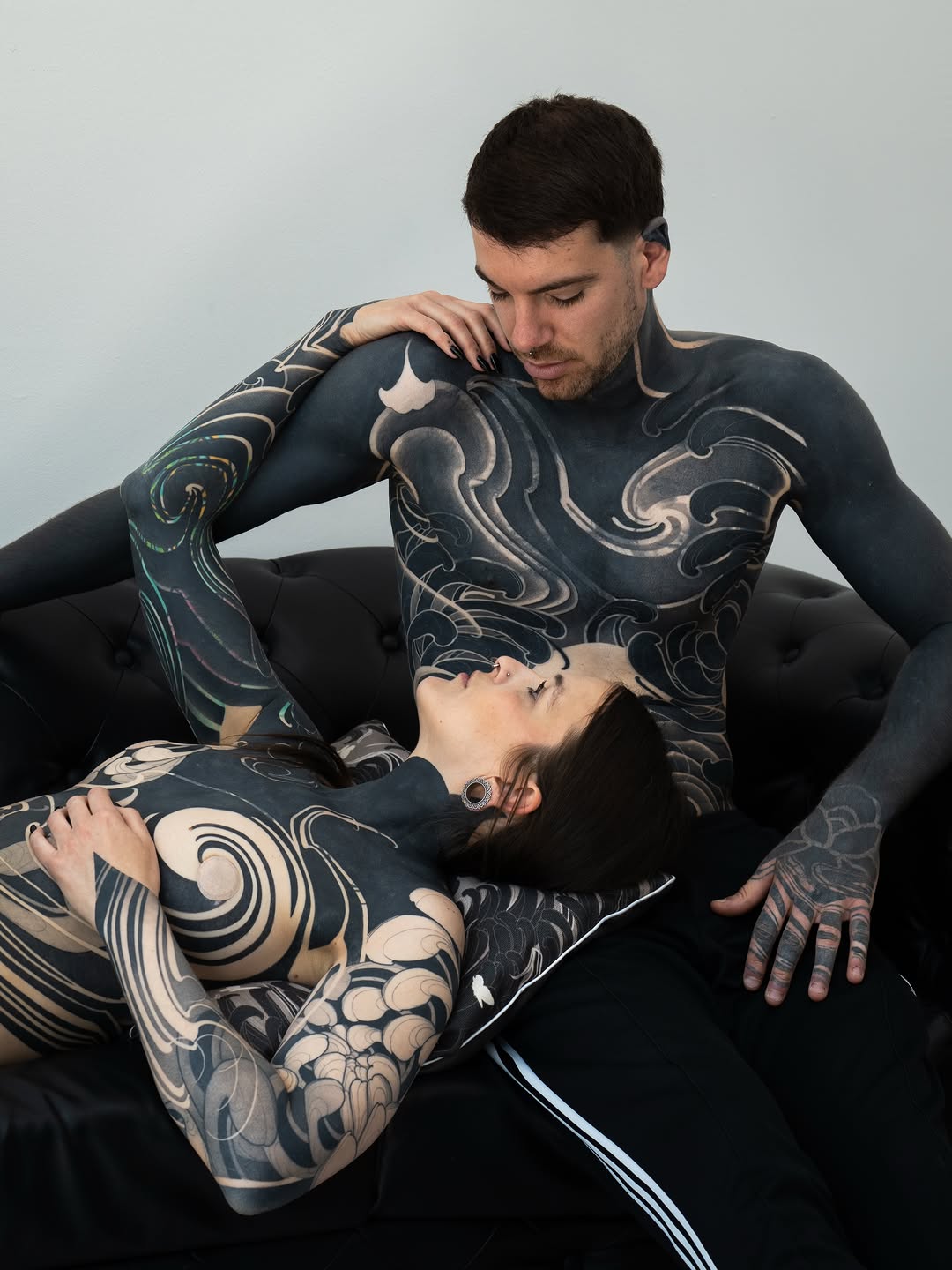 Two heavily tattooed people in black clothing pose together, displaying intricate monochromatic designs covering their arms, legs, and torsos in swirling, ornamental patterns.