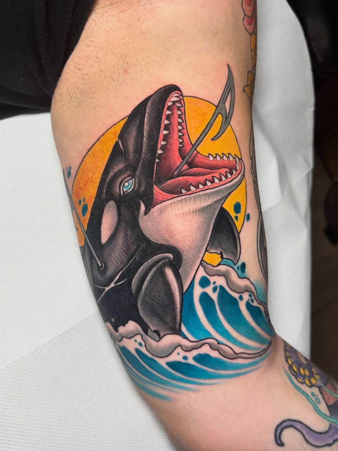Orca whale tattoo on upper arm with open mouth revealing red interior, orange sun background, and blue ocean waves below.