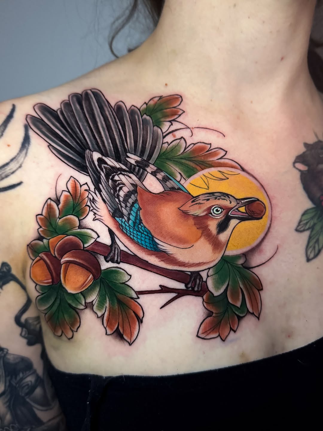 Colorful bird tattoo with black wings, orange and yellow body, and blue wing detail, surrounded by orange and red flowers with green leaves on the chest.