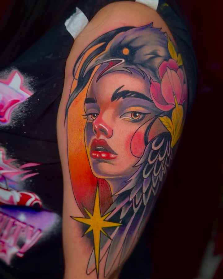 Colorful tattoo on upper arm featuring a woman's face with bird wings, flowers, and a star in orange, pink, yellow, and purple tones.
