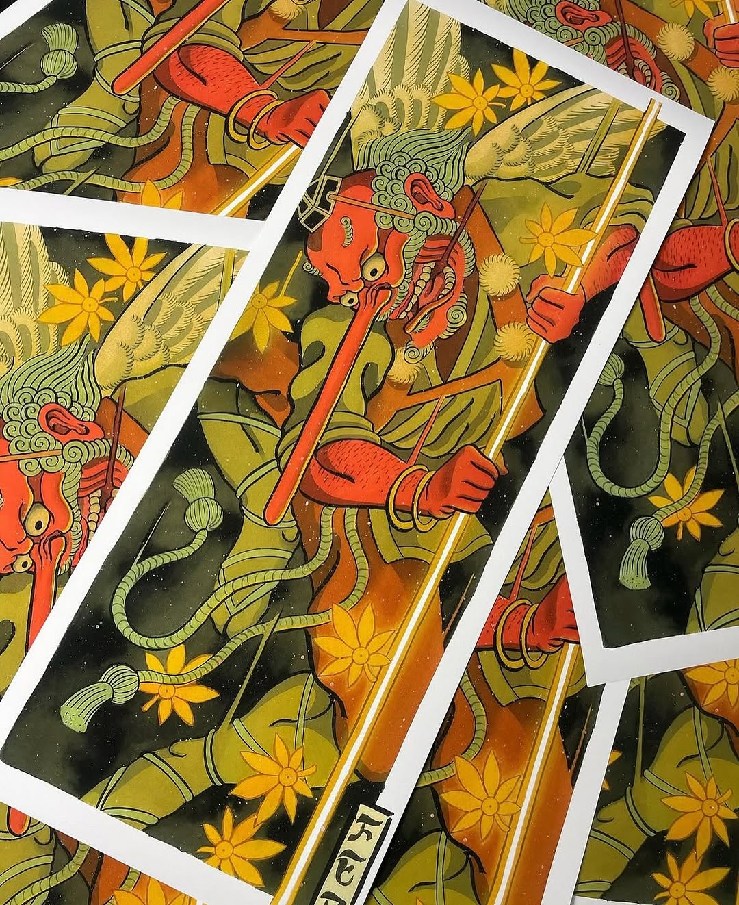 Multiple printed reference sheets displaying a red demon mask with ornate details, yellow flowers, and green foliage on dark backgrounds in warm earth tones.
