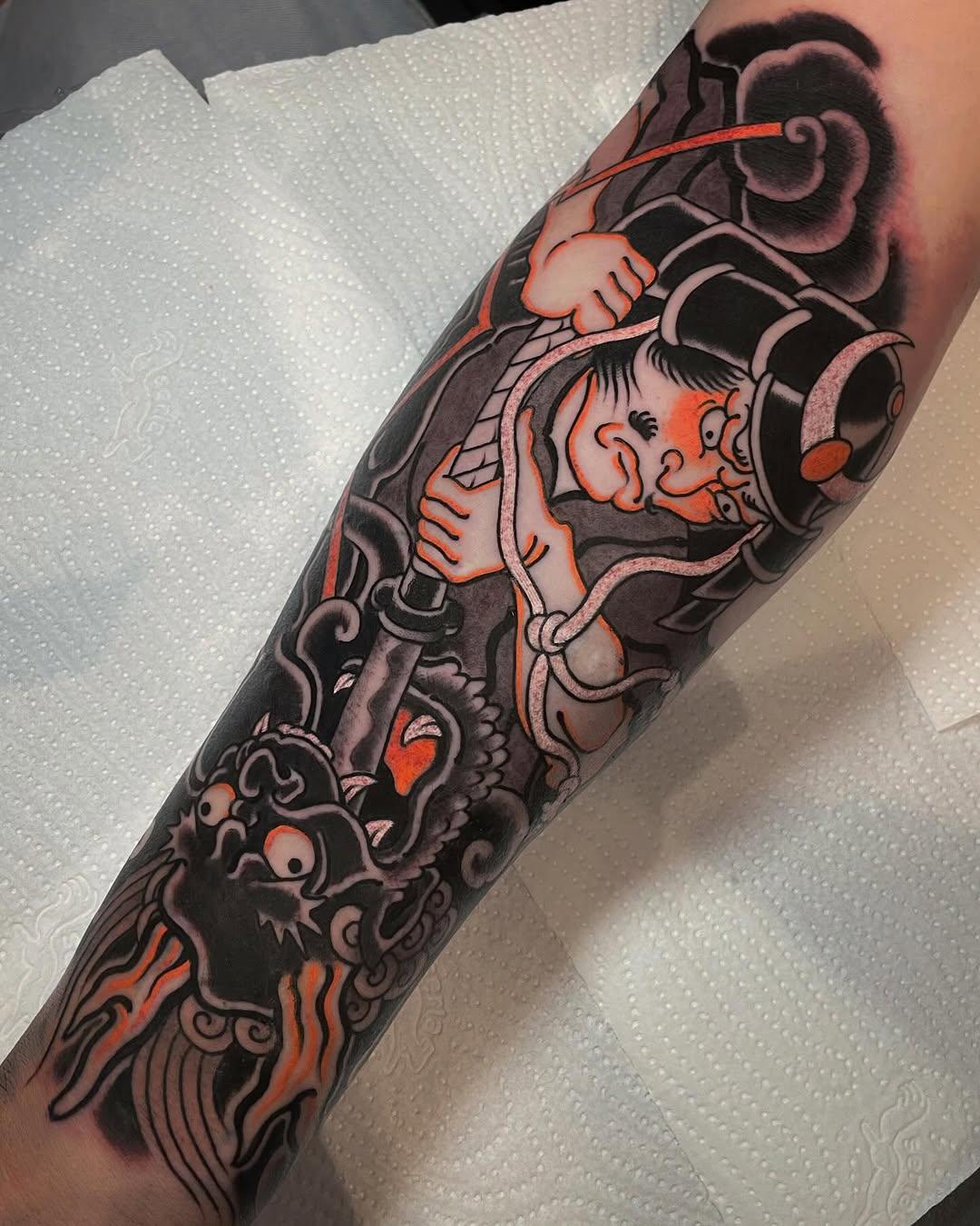 Full-sleeve tattoo on arm featuring ornate mechanical gears, demon faces, and decorative scrollwork in black, grey, and orange ink.