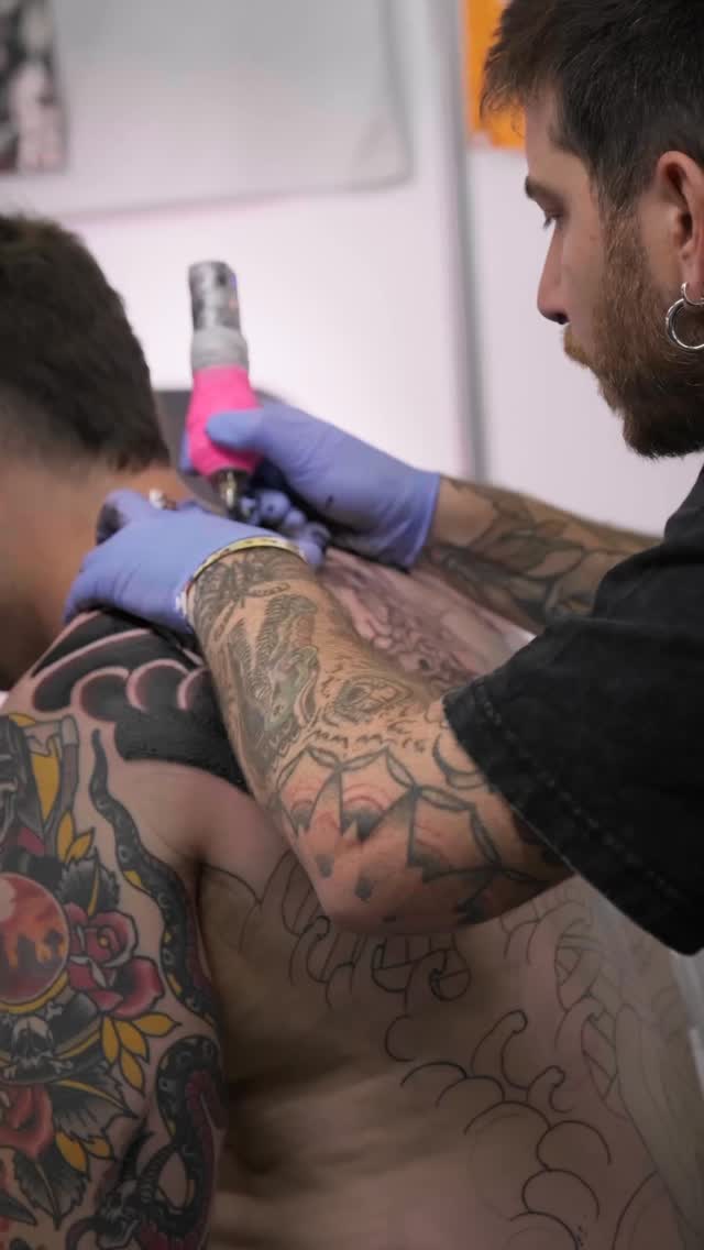 Tattoo artist applying pink and blue ink to client's shoulder and upper arm featuring existing colorful floral and decorative designs.
