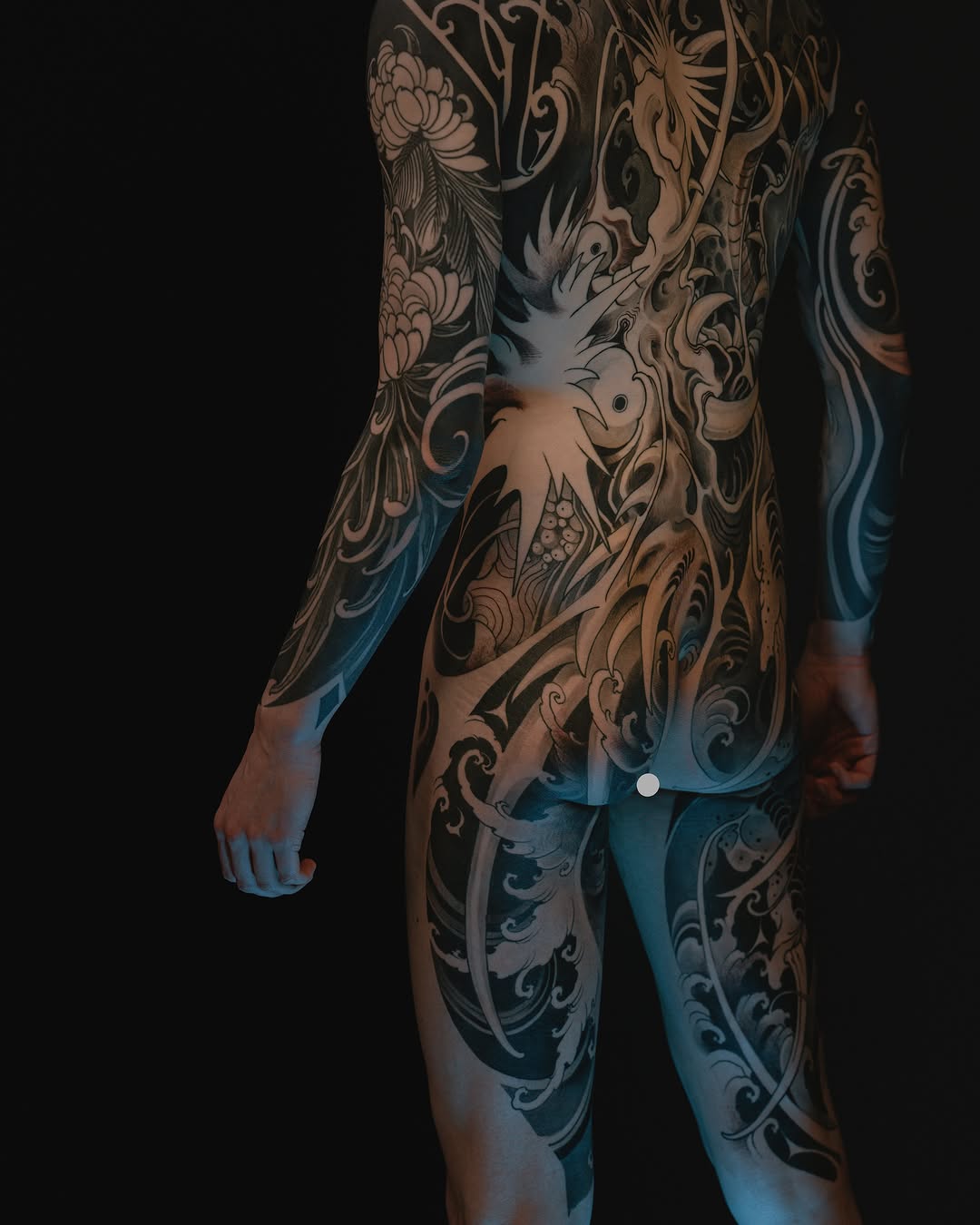 Full-body tattoo featuring ornate scrollwork, floral motifs, and a central phoenix design in black, tan, and blue tones covering the torso, arms, and legs.