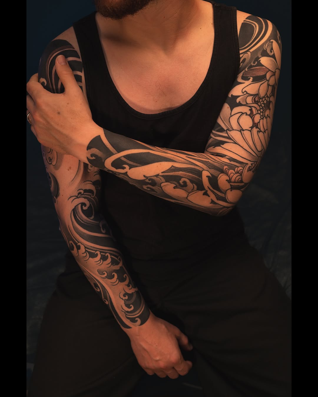 Man displaying full-sleeve tattoos with ornamental scrollwork, floral motifs, and curved designs in black ink on both arms.