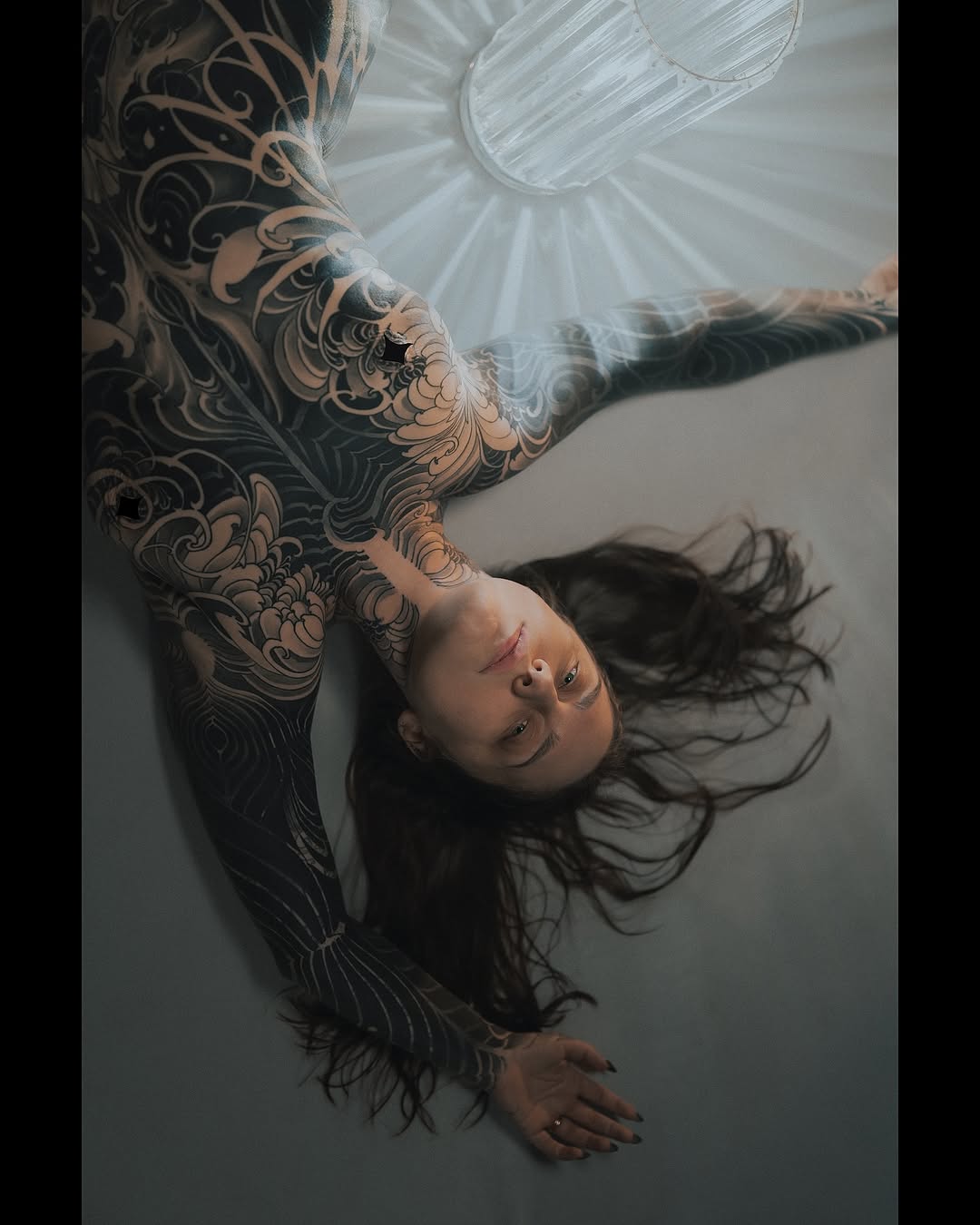 Overhead view of a person with extensive dark ornamental patterned tattoos covering their arm and leg, lying on a light background.