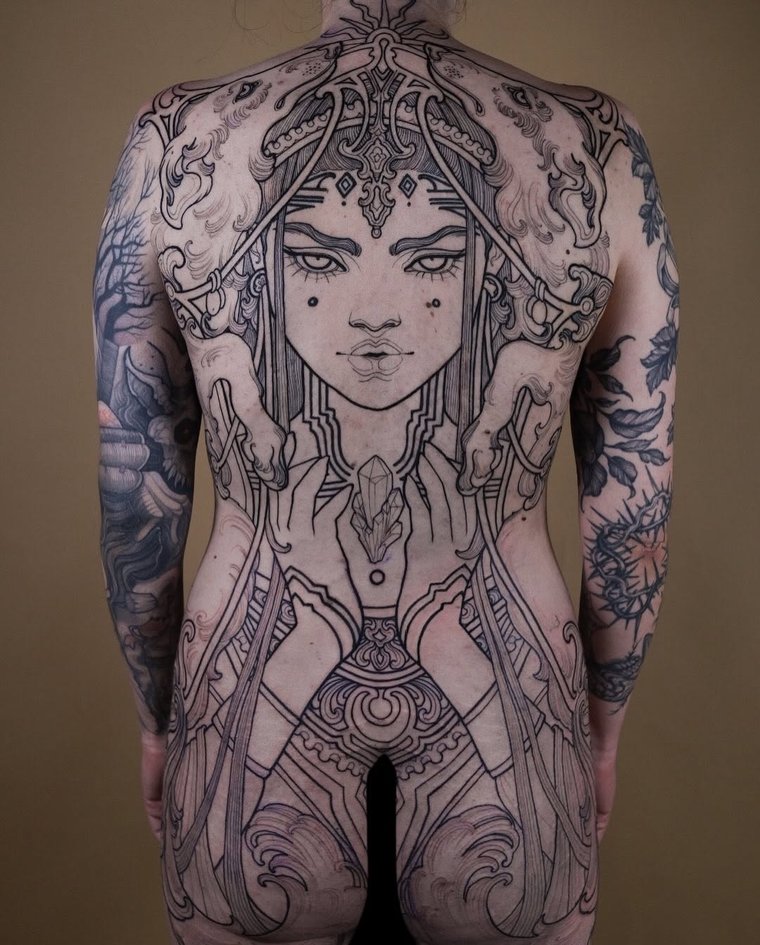 Back tattoo featuring a centered female face with ornate decorative headdress and symmetrical flowing patterns extending down the spine and sides in black ink.