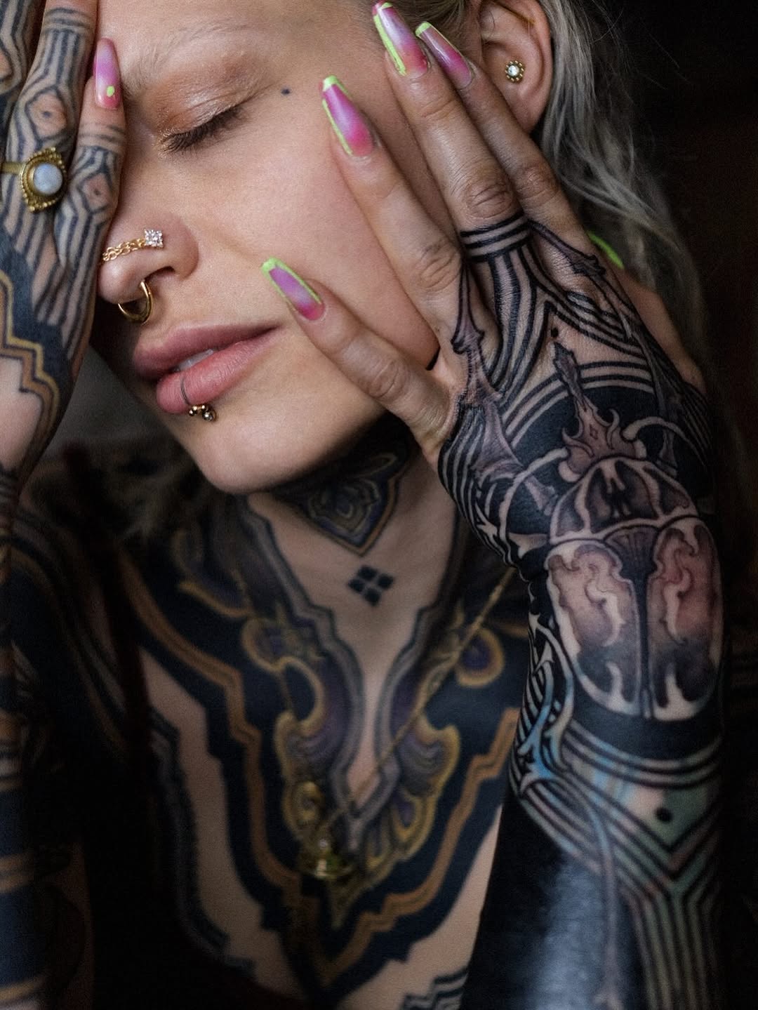 Tattooed person with extensive black and gold chest and arm designs displays purple and neon green ombre nails while posing with hands near face.