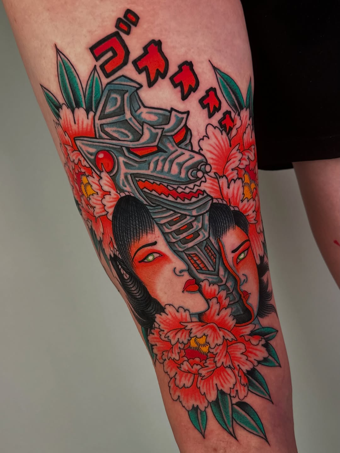 Colorful leg tattoo featuring a woman's face with red overlay, mechanical skull headdress, red flowers, green leaves, and red text on the upper thigh.