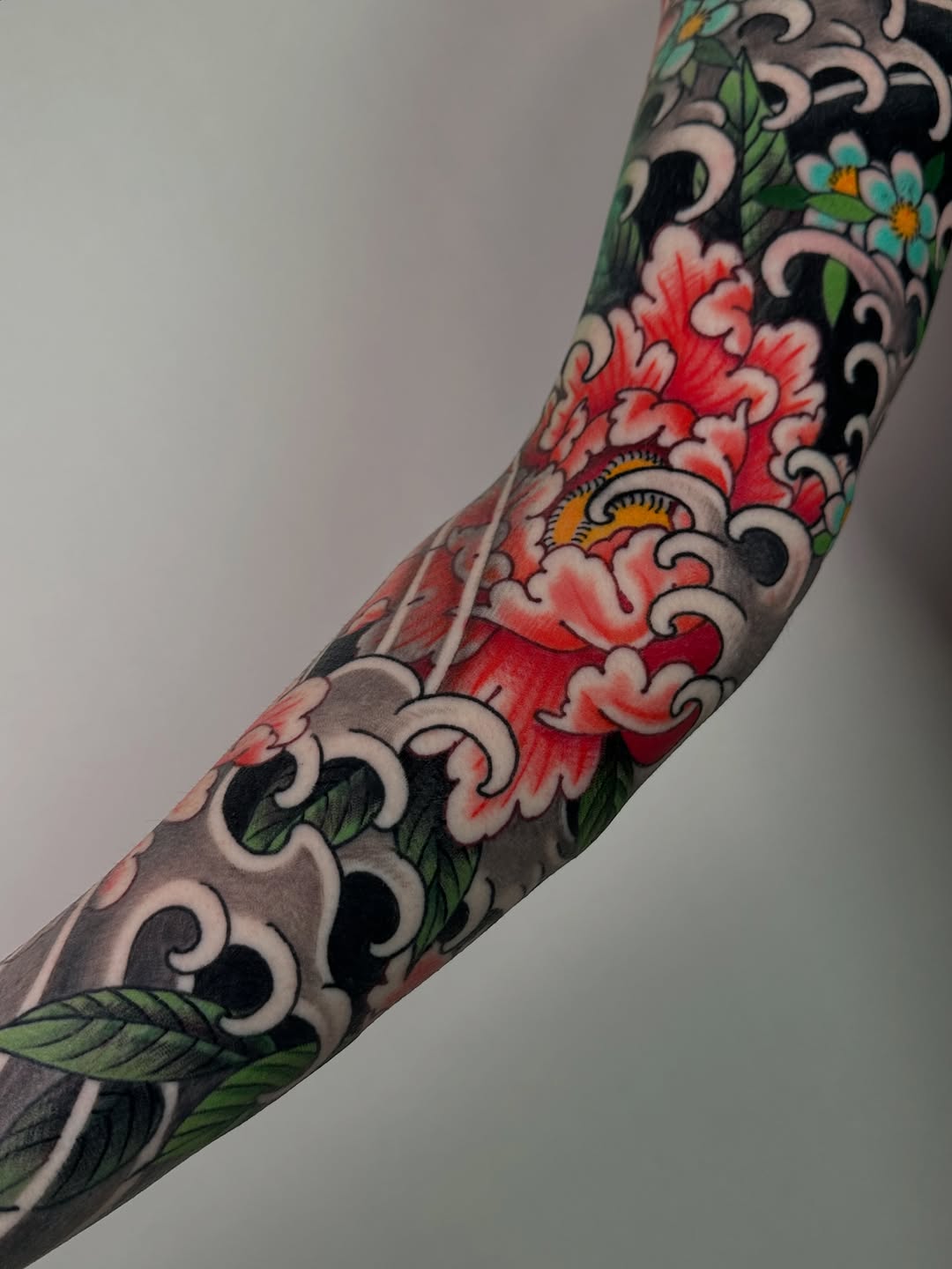 Colorful arm sleeve tattoo featuring red and blue flowers with green leaves, ornamental scrollwork, and eye motifs in black and cream on gray skin.
