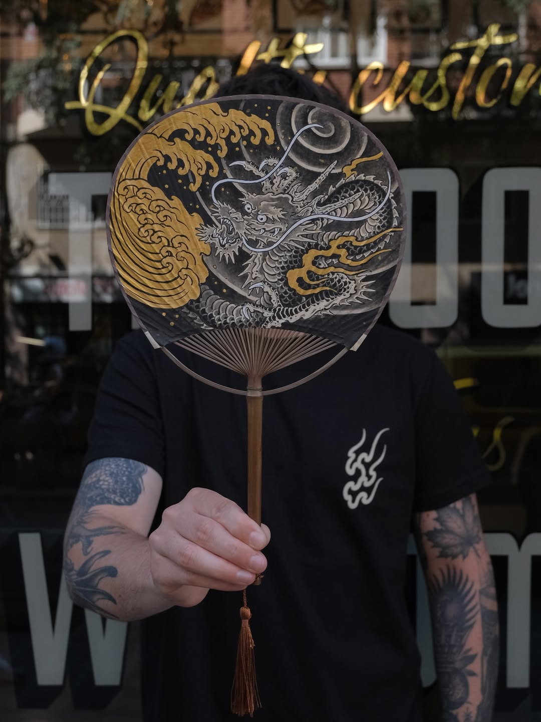 Person with tattooed arms holding a decorative fan with gold and silver dragon and wave designs against a storefront window.