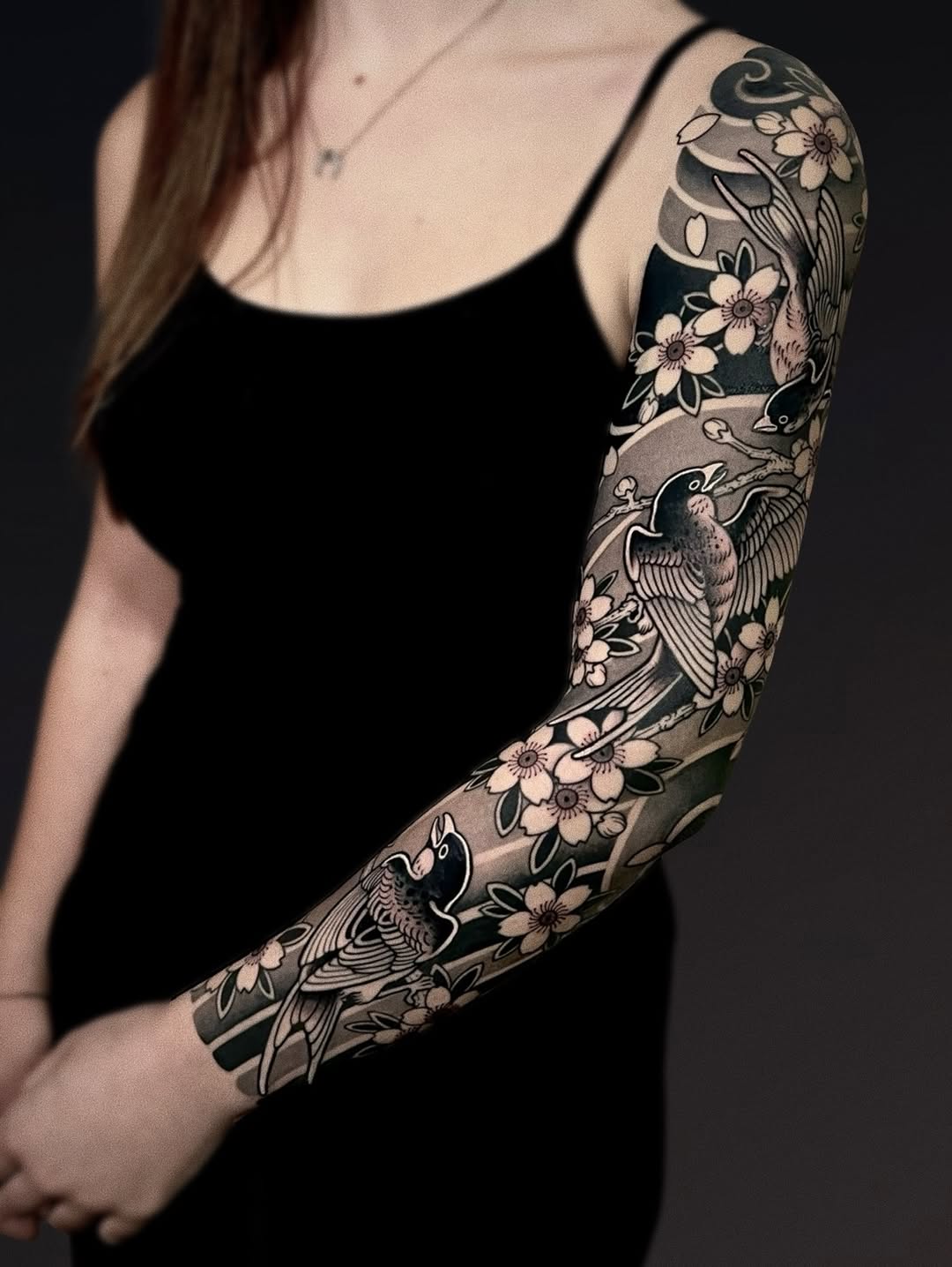 Full arm sleeve tattoo featuring birds and flowers in black ink with grey shading on a woman's right arm against a dark background.