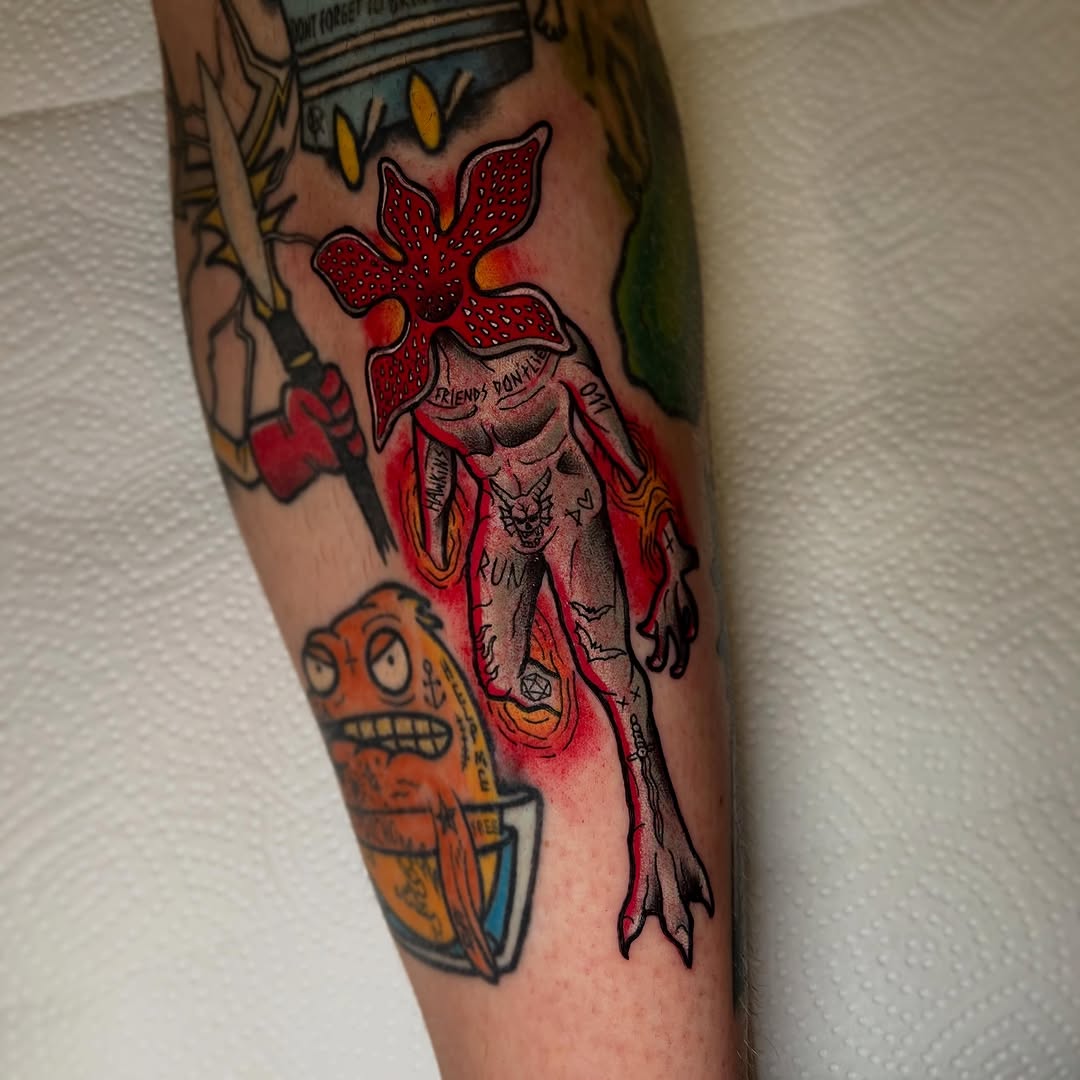 Colorful tattoo on an arm featuring a red flower with a skeletal figure, text, and an orange character with geometric patterns below.