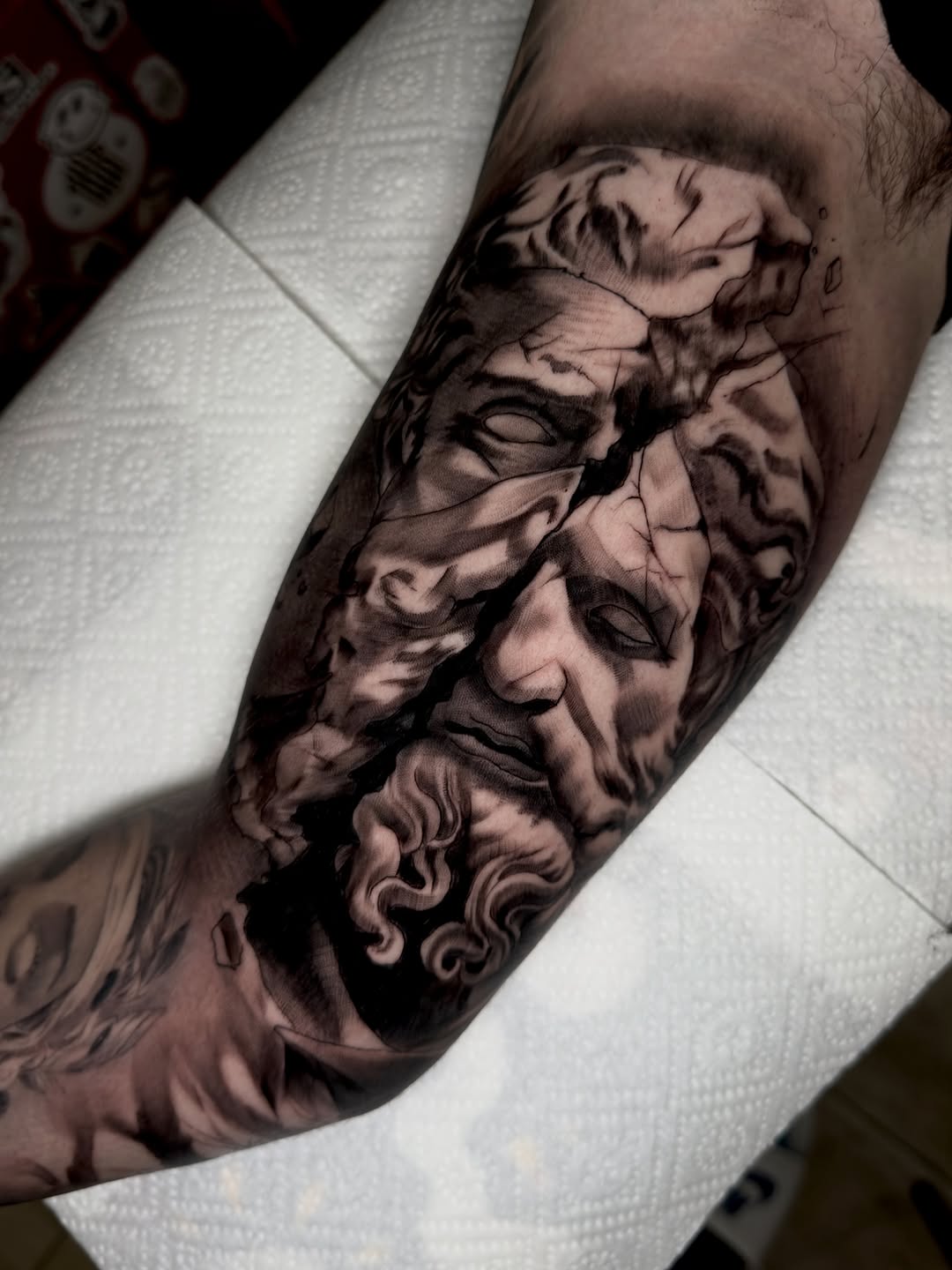 Detailed sculptural male face and cherub tattoo on forearm in black and gray ink with extensive shading and dimensional detail.