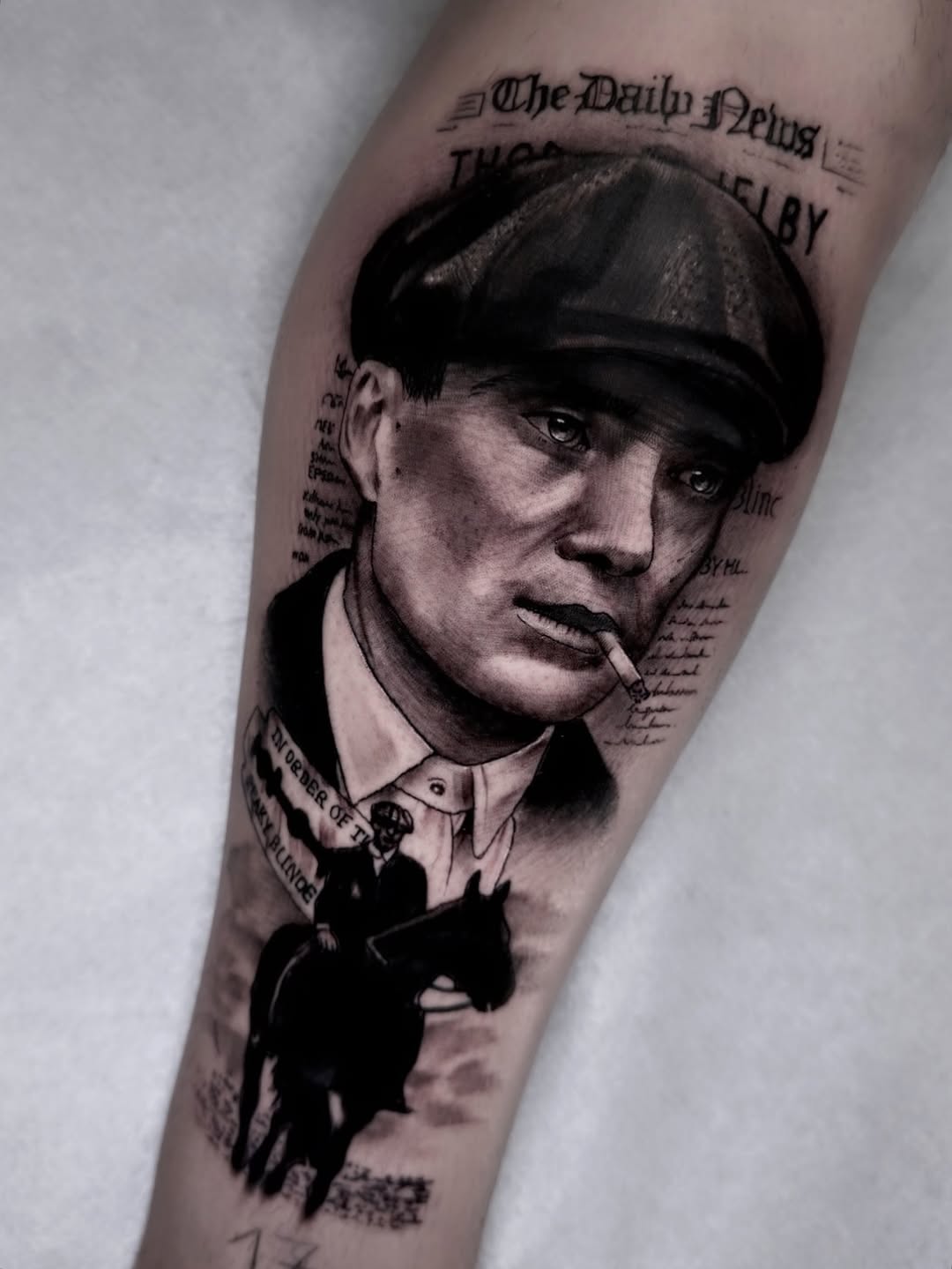 Portrait tattoo on forearm featuring a man in a flat cap with cigarette, newspaper clippings, and silhouetted figures in black ink.