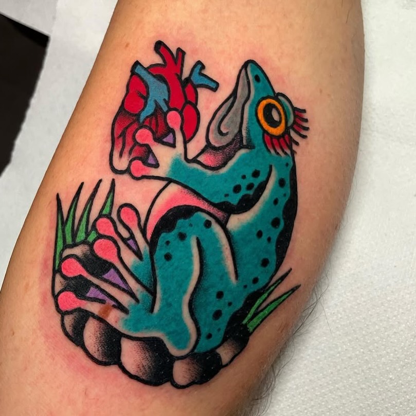 Colorful tattoo on an arm featuring a turquoise axolotl holding a red anatomical heart, with pink flowers and green plants below, outlined in black.