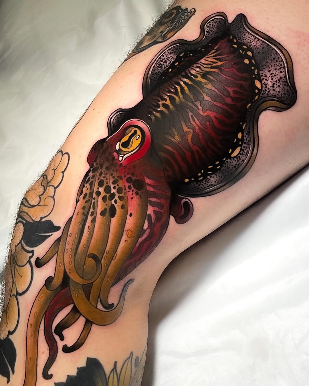 Detailed octopus tattoo on upper arm with tentacles extending downward, featuring warm orange and red tones with black outlines and textured shading.