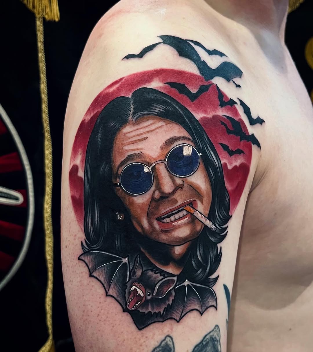 Portrait tattoo on upper arm featuring a person with long black hair, round sunglasses, and cigarette, surrounded by bats and a red circular background.