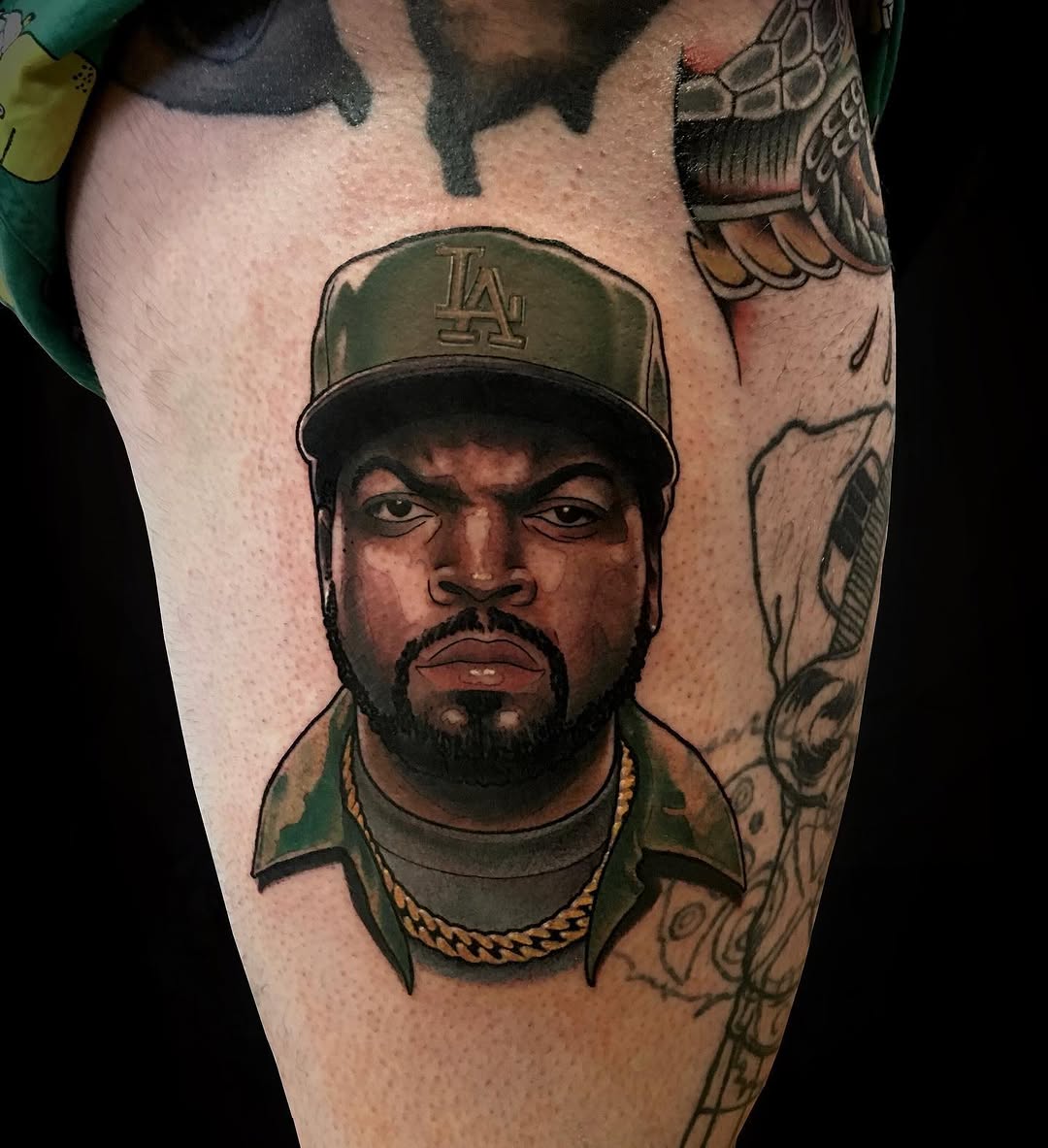 Portrait tattoo on thigh of a person wearing an LA baseball cap and gold chain necklace, rendered in brown, green, and black ink.