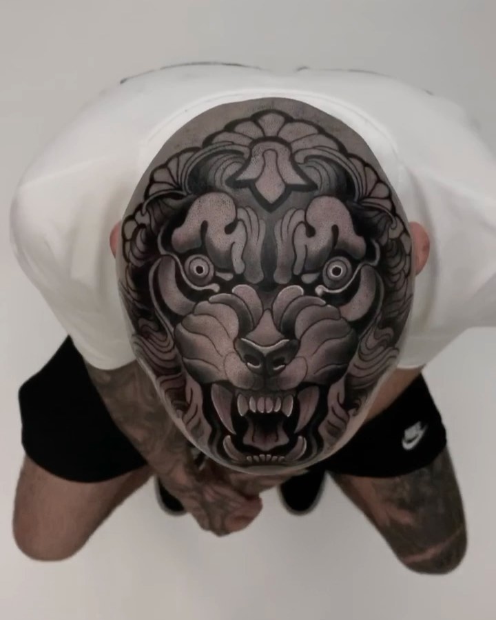 Overhead view of a detailed demon or oni mask tattoo on a person's scalp, featuring intricate shading in grays and blacks with prominent eyes and bared teeth.