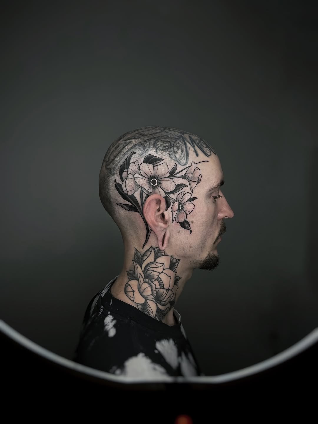 Man shown in profile with extensive floral tattoos on head, neck, and shoulders in black ink with pink ear detail against dark background.