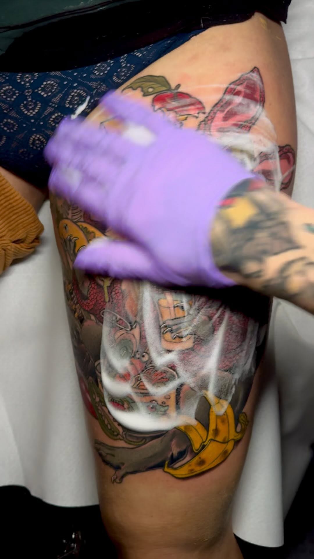 Colorful leg tattoo featuring fish, bananas, and architectural elements in red, yellow, green, and orange on the thigh, shown with purple wrapping.