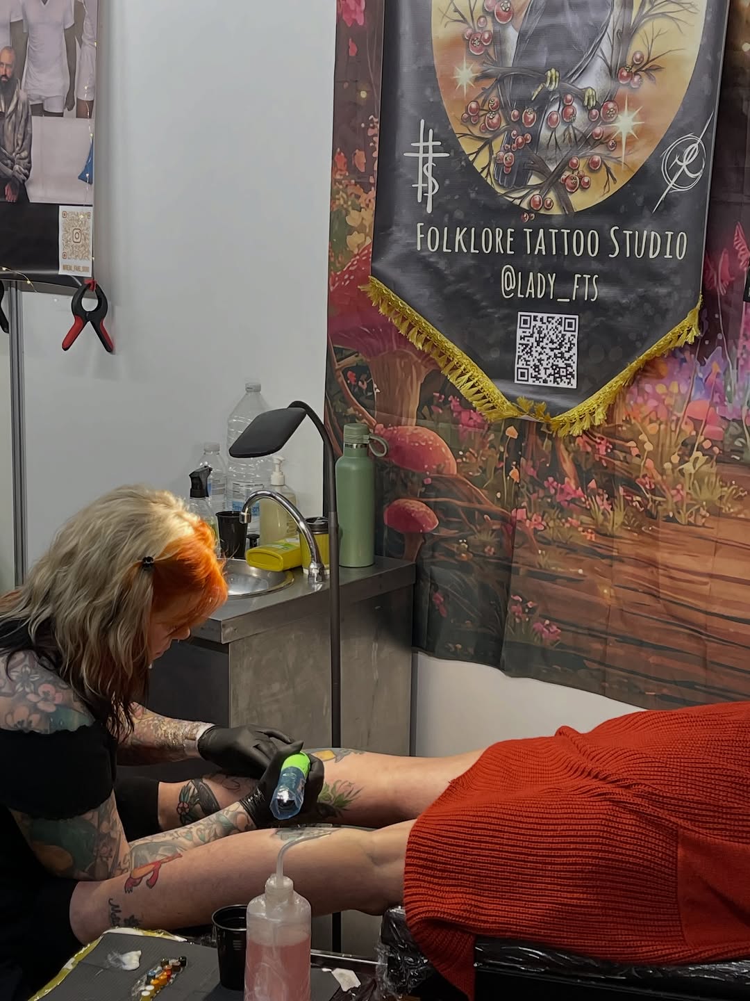 Tattoo artist with orange-tinted hair applies a colorful design to a client's arm wearing a red sweater at Folklore Tattoo Studio.