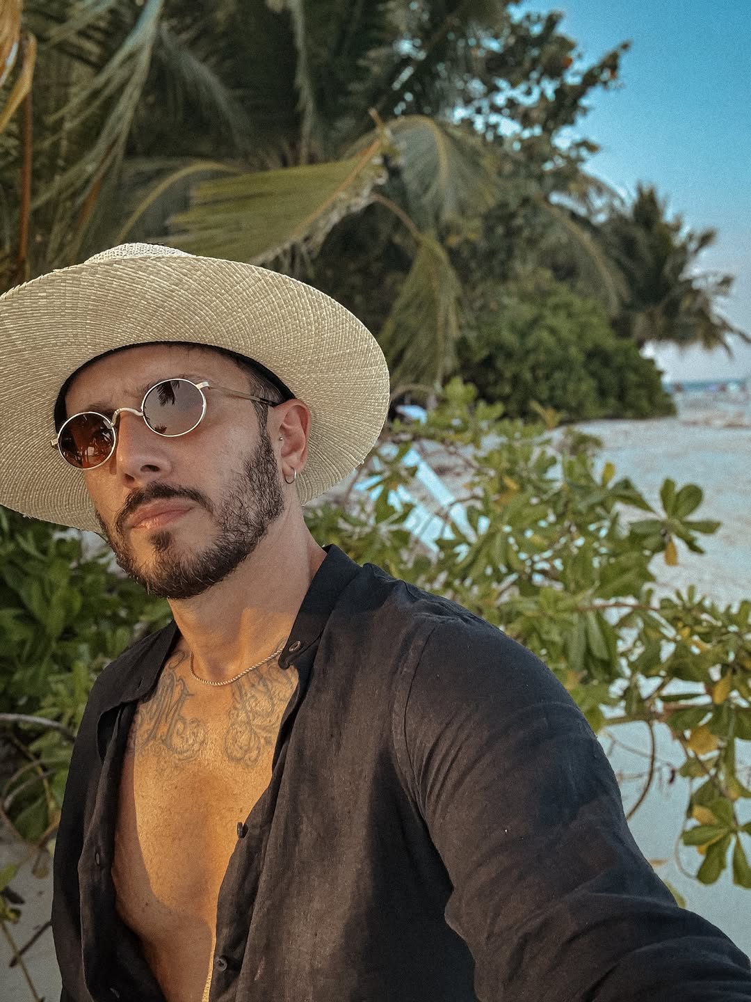 Man wearing a detailed chest tattoo with ornamental designs in warm tones, photographed outdoors wearing a straw hat and sunglasses.