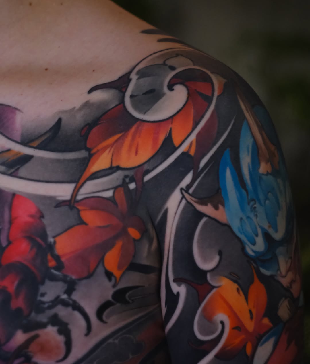 Colorful bird tattoo on shoulder featuring orange, red, and blue wing details with decorative curved line work and dark shading.