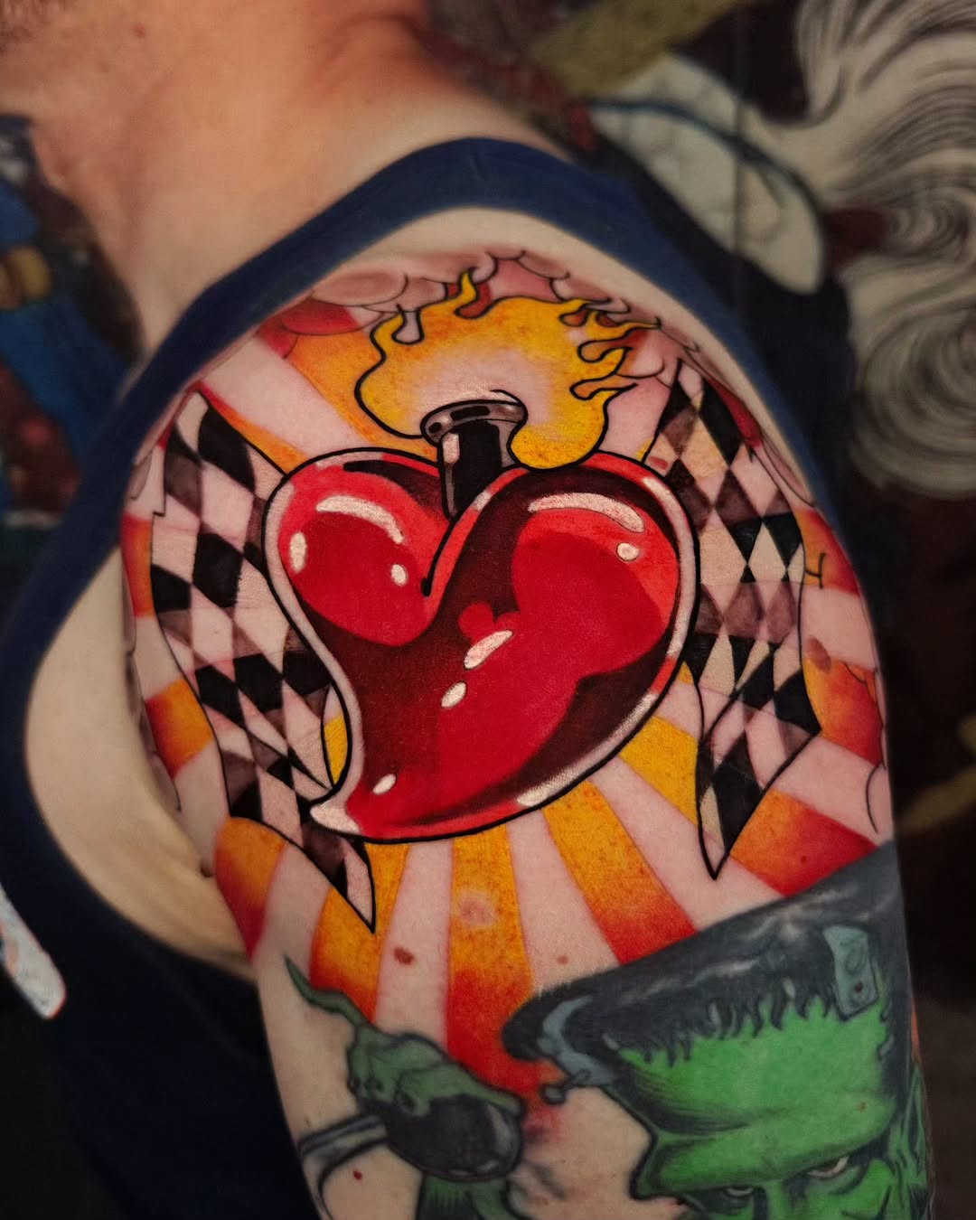 Sacred heart tattoo with flames, harlequin pattern background, and green element below on upper arm in red, yellow, orange, and black.