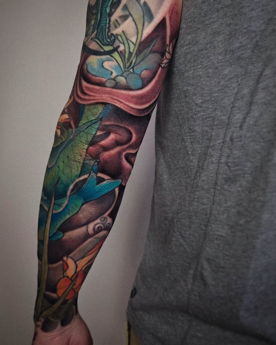 Colorful full-length arm tattoo featuring mythological figures, blue and teal fabrics, burgundy draping, and botanical elements on the forearm and upper arm.