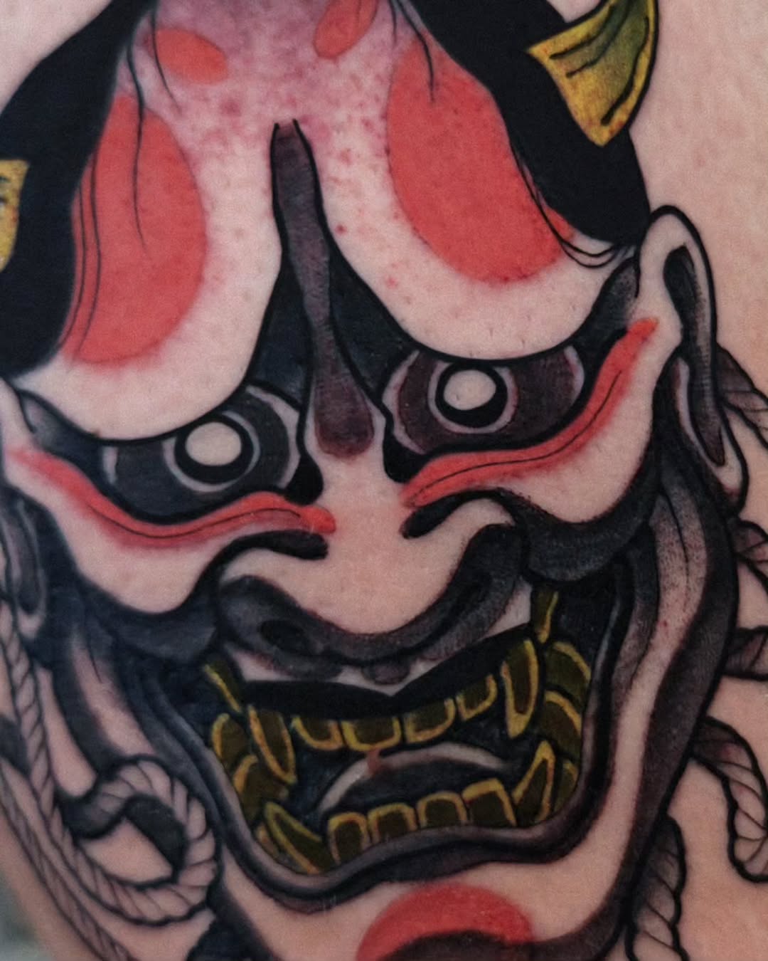 Demon or oni mask tattoo with red and black coloring, gold accents, and detailed facial features on skin.