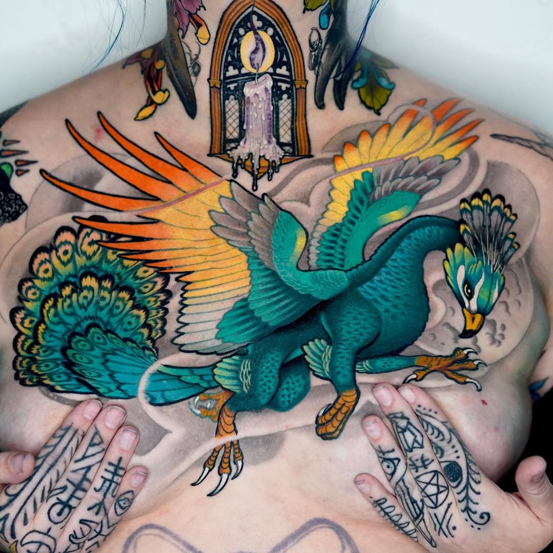 Colorful chest tattoo of a teal and orange winged peacock with extended feathers, flanked by a candle window above and decorative hand tattoos below.
