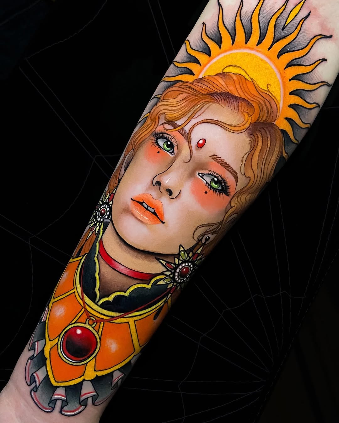 Colorful arm tattoo featuring a woman with wavy auburn hair, green eyes, orange sun halo, jeweled ornaments, and golden clothing against black background.