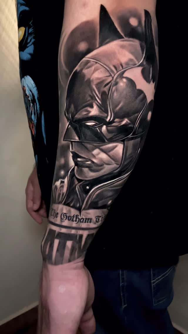 Batman cowl portrait tattoo on upper arm with "The Gotham" text band, rendered in black and gray ink with shading.