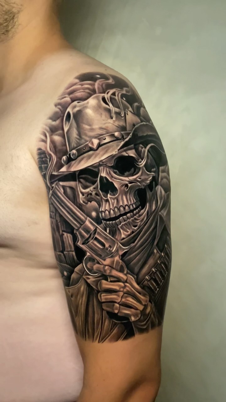 Detailed skull tattoo on upper arm wearing a leather strap and holding a revolver, rendered in grayscale with shading on pale skin.