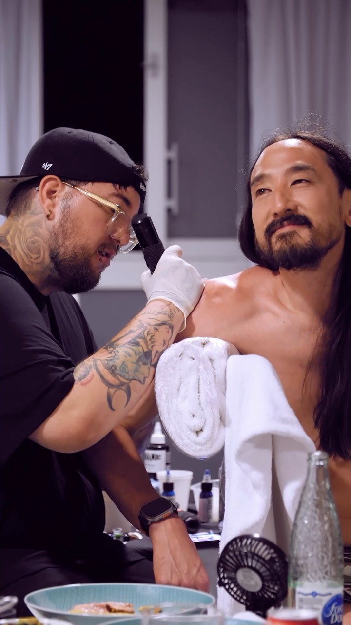 Tattoo artist applies makeup or applies detail work to client's arm featuring a large black tribal design during a professional session.