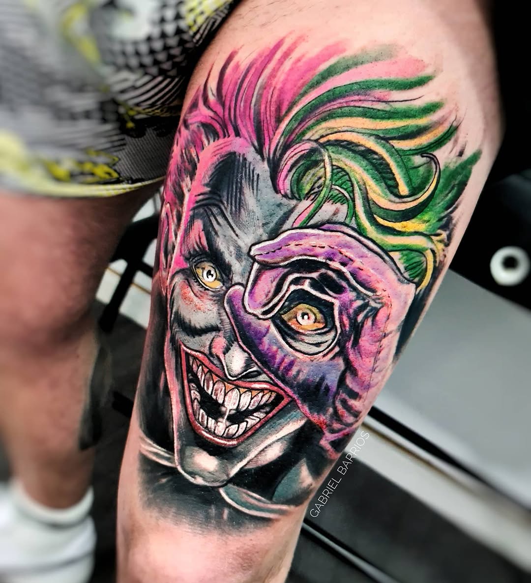 Colorful jester or demon face tattoo on upper arm featuring pink, green, gold, and purple with exaggerated features and sharp teeth.
