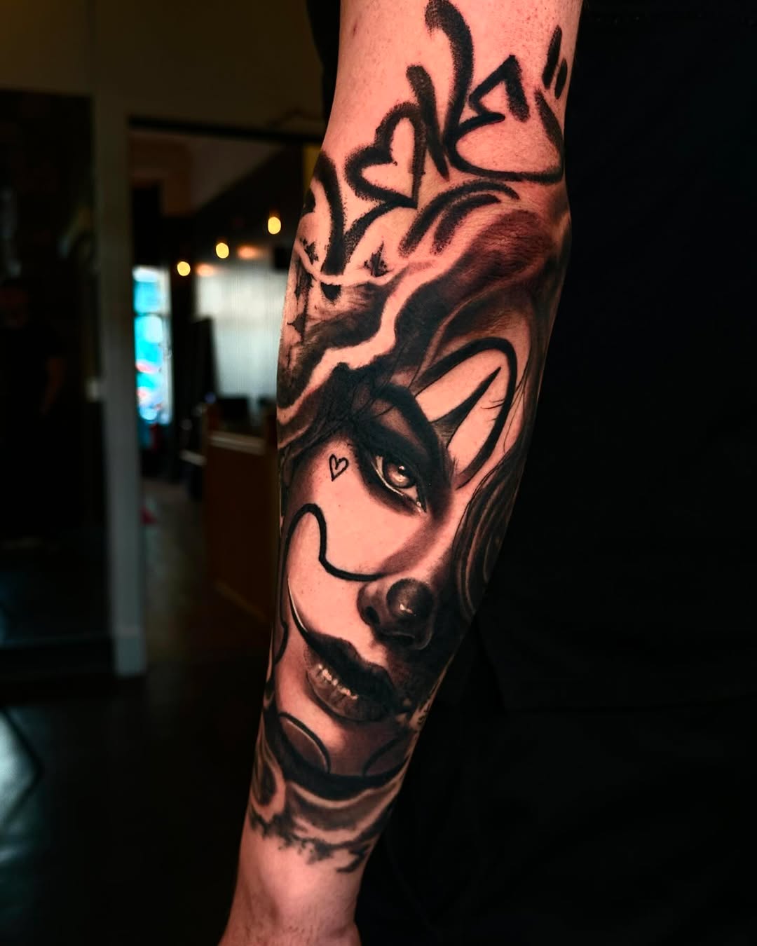 Demon face tattoo on the forearm with detailed shading, sharp facial features, horns, and a small heart marking near the eye in black ink.