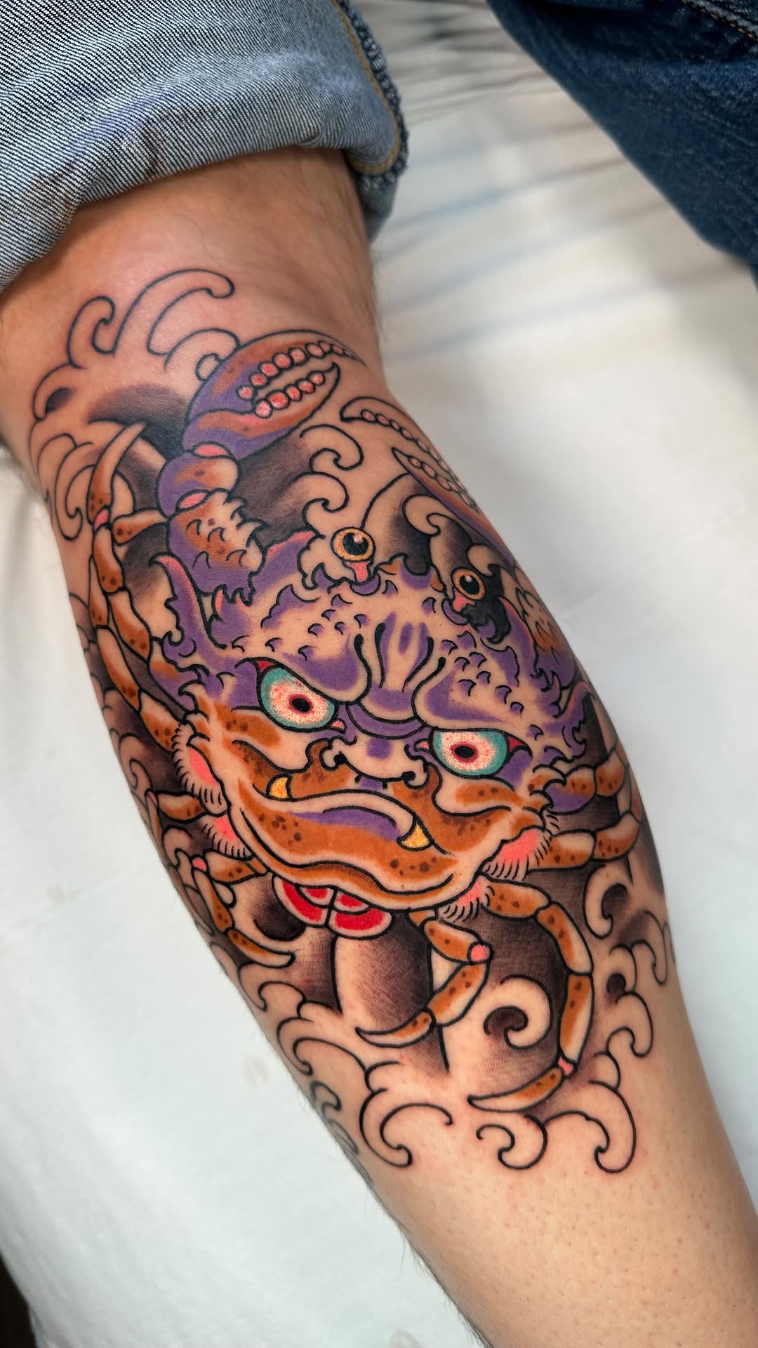Colorful oni mask tattoo on the forearm featuring orange and purple tones with ornate swirling details, red mouth, and piercing eyes.