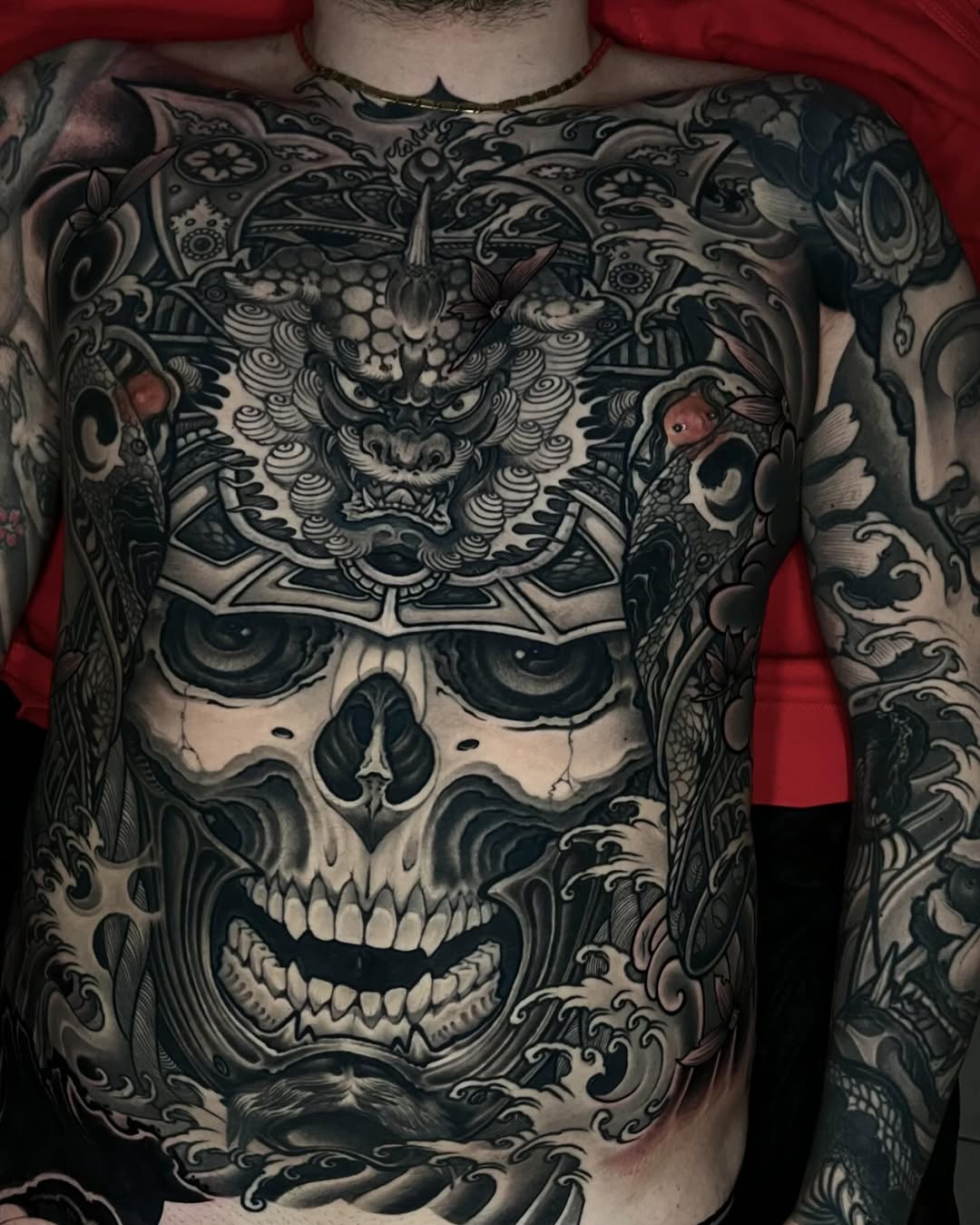 Large chest and sleeve tattoo featuring an ornate demon mask above a skull with decorative scrollwork and floral elements in black and grey ink.