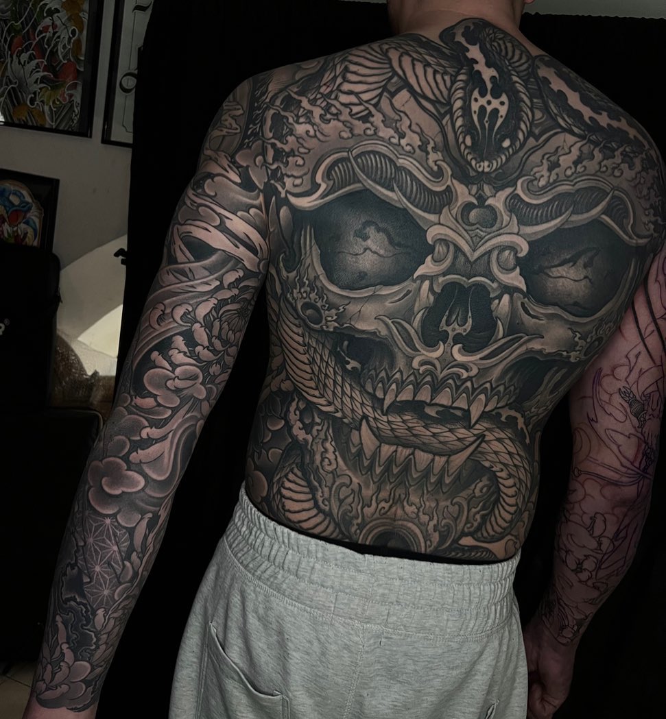 Shirtless man displaying full back and sleeve tattoos featuring a large ornate skull with decorative flourishes and floral patterns in black and grey ink.