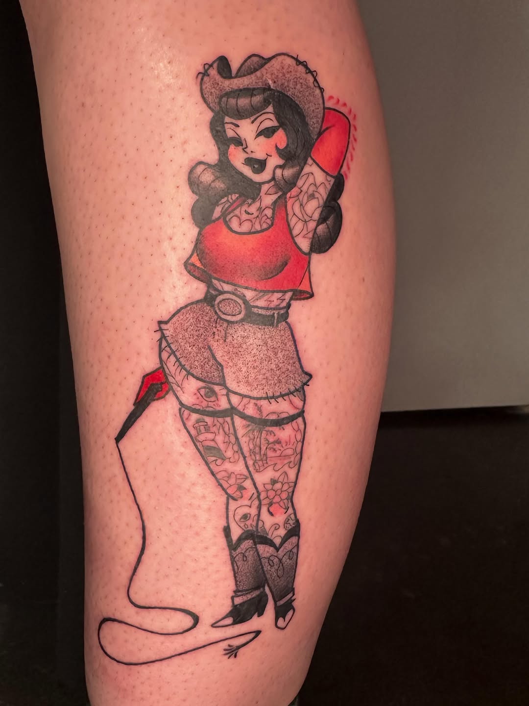Colorful tattoo on calf featuring a pinup-style woman in red clothing with tattooed legs, holding a whip, with black outlines and red accents.