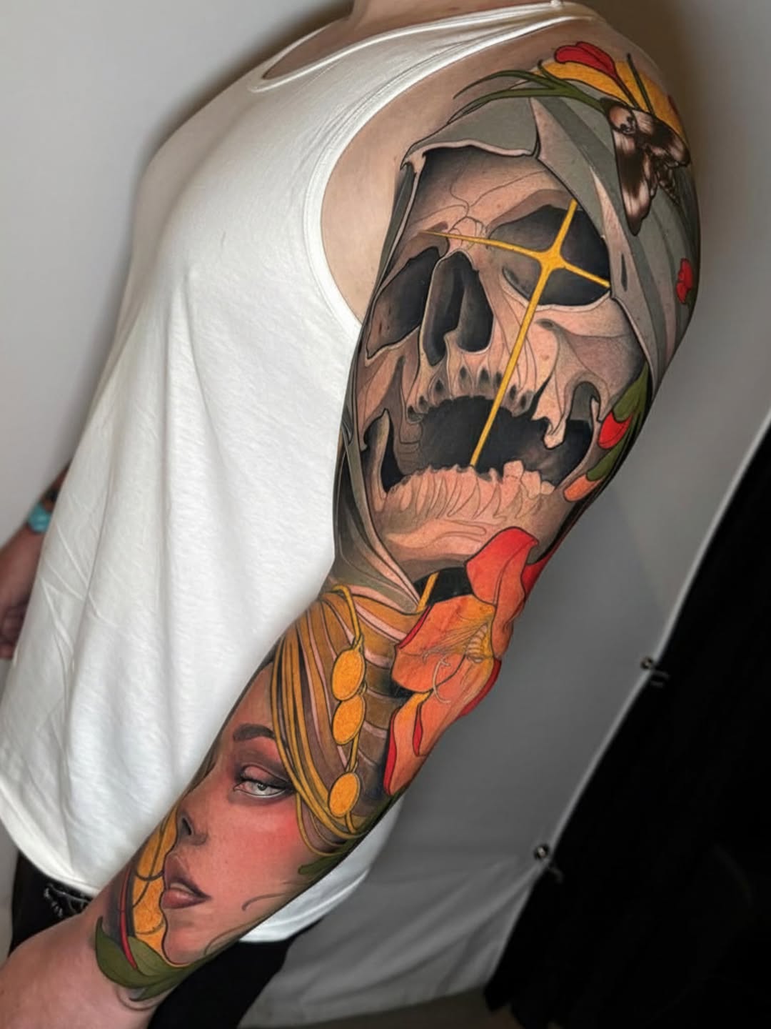 Colorful sleeve tattoo featuring a skull with flowers, a female face in profile, and anatomical elements in red, orange, yellow, and green tones.